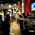 Bancone del bar con davanti due drink e il conto e con sfondo persone al bancone